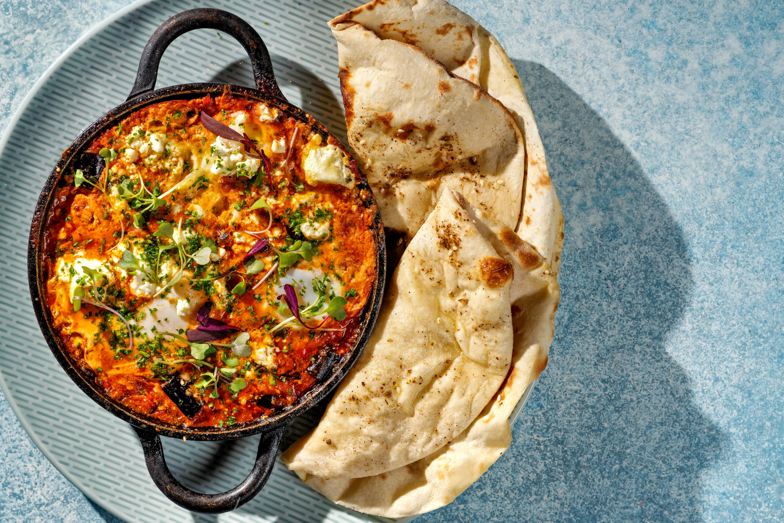 Shakshuka dish in a cast iron , two-handled skillet served with a large crescent-shaped pita bread. Naxos Taverna